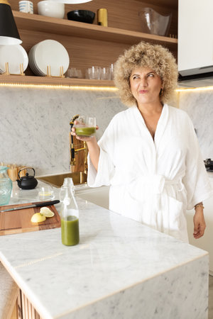 Curly-Haired Woman Enjoying Green Juice in Modern Kitchenの写真素材