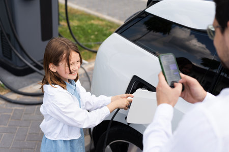 Smiling Girl Charging Electric Car with Parental Supervision at Charging Stationの写真素材