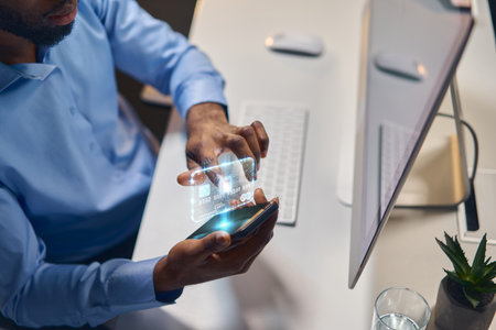 Man Using Smartphone With Holographic Credit Card Interface Near Modern Desktop Computerの写真素材