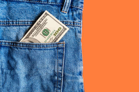 Money in pocket. One hundred dollar bill in a denim jeans pocket on a orange background. Copy space. Concept of earning and saving money. American moneyの写真素材