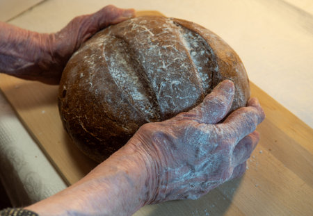 Black fresh round rye bread in the hands of an elderly woman on a kitchen board. Bread in flour....