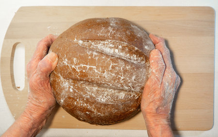 Black fresh round rye bread in the hands of an elderly man on the kitchen board.  Bread in flour