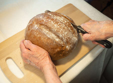 Elderly woman's hands cut round rye bread in flour on kitchen boardの写真素材