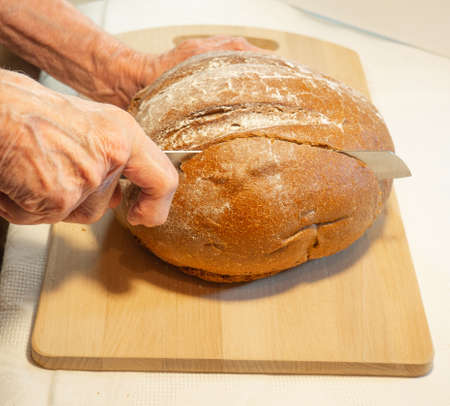 Elderly woman's hands cut round rye bread in flour on kitchen boardの写真素材