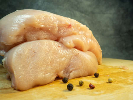 Chicken breast fillet pieces on a cutting board. Still life on a black background. Multi-colored peppercorns around a piece of meat. Cooking chicken. Raw meat.の写真素材