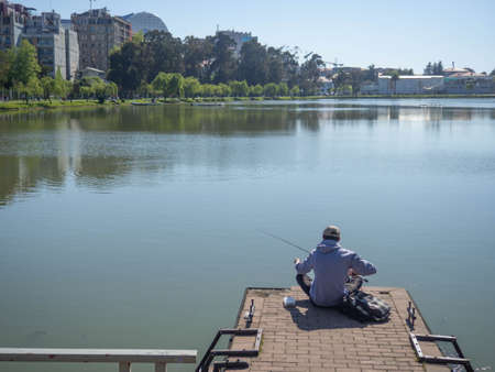 A fisherman with a spinning rod sits on the pier of the lake. Quiet pastime. sports and recreation. hobby. Waiting for a biteの写真素材