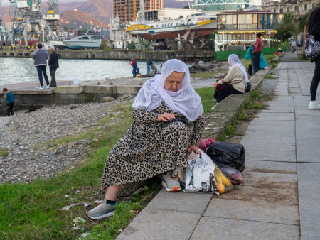 Batumi, Georgia. 10/24/2022 An elderly woman in a headscarf looks at the phone. old lady and modern technology. A woman in a headscarf and with bags on the shore.のeditorial素材