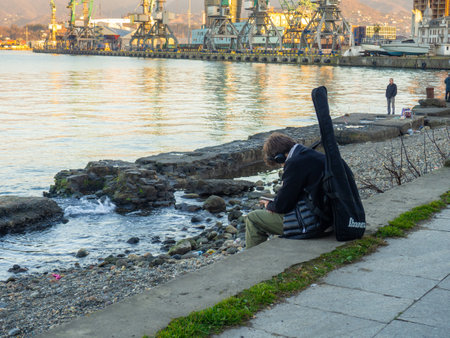 Batumi, Georgia. 21/01/2023 A guy with a guitar in a case is sitting on the seashore. The concept of creative thinking. Musician at sea. Listening to music and reading.のeditorial素材