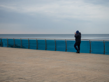 A man walks along the bridge along the seashore. Walking in the resort in the winter season.  man in the hood speaks on the phone. Coast of the Black Sea. Batumiの写真素材