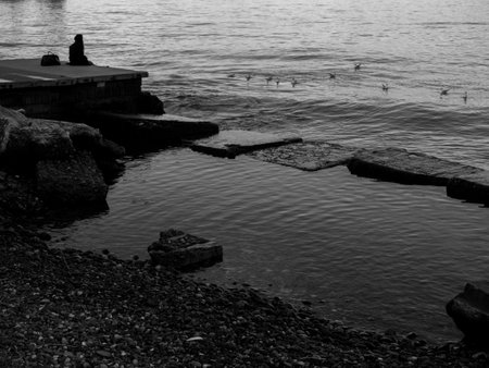 Black and white photography. A lonely girl sits on a pier in the port. Sitting on the edge of the water.   The concept of loneliness.の写真素材