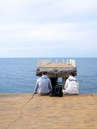 Two sit on the pier with their legs dangling. Two guys at sea. Friends met and talk on the old pier. Destroyed infrastructure at the resort. The concept of privacyの写真素材