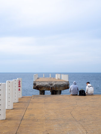Batumi, Georgia. 03.11.2023  Two sit on the pier with their legs dangling. Two guys at sea. Friends met and talk on the old pier. Destroyed infrastructure at the resort. The concept of privacyのeditorial素材
