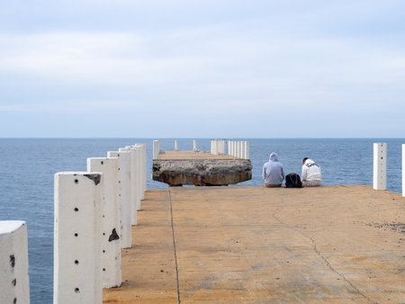 Batumi, Georgia. 03.11.2023  Two sit on the pier with their legs dangling. Two guys at sea. Friends met and talk on the old pier. Destroyed infrastructure at the resort. The concept of privacyのeditorial素材
