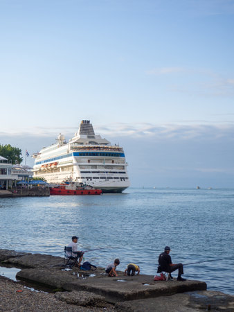 Batumi, Georgia. 07.31.2023  A fisherman is fishing on the pier with a cruise ship in the background. International seaport. Batumi. Caught fish in the sea with a lineのeditorial素材