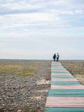 A couple holding hands walks along the path to the sea. Lovers against the backdrop of the sea. Silhouettes of people. Road made of wooden palletsの写真素材