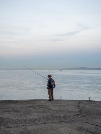 Batumi, Georgia. 12.04.2023 Lonely fisherman with a fishing rod on the pier in the evening. A man is sitting in the port area. Loneliness concept. Port city in winter. At duskのeditorial素材