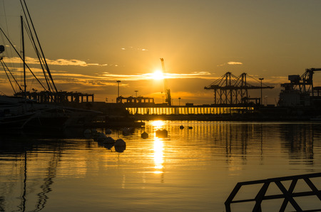 sunrise at the Valencia harbor, some clouds sun rises between docked sailboats and cargo port cranes orange red colors in the sky and water reflectionの写真素材