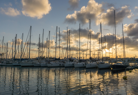 Afternoon sunset between clouds reflected over calm sea water, blue and orange colors. Sailboats with their masts in row at the marinaの写真素材