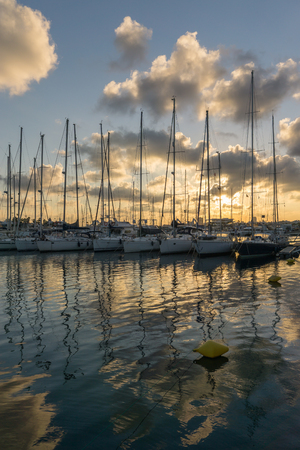 Afternoon sunset between clouds reflected over calm sea water, blue and orange colors. Sailboats in row at the marina, vertical viewの写真素材