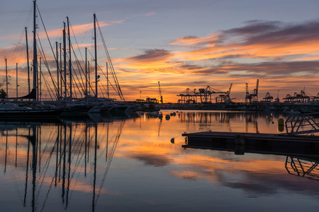 Twilight at harbor sunrises silhouettes of docked sailboats and cargo port cranes red orange colors in the sky and water reflection horizonの写真素材