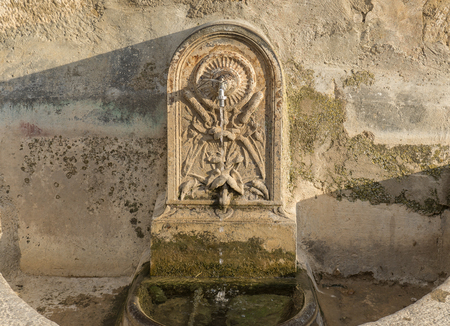 Ancient water fountain carved in stone, with restored modern faucet water falling, side illuminated by the sunの写真素材