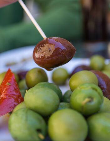 Extreme Close-up of Black Olive caught with a toothpick from a spiced olives plate on tapas bar table a typical Spanish black and green split variety in detail. Vertical viewの写真素材