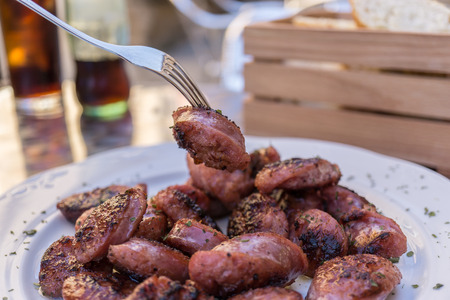 Piece of sausage caught with fork. Grilled sausages from Aragon Teruel Spain, Cut into pieces, typical tapas dish. Close-up. Blurred background basket of bread and sodaの写真素材