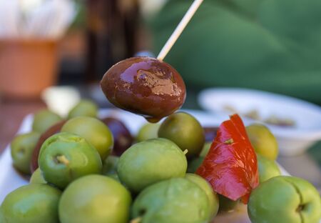 Close-up of Black Olive caught with a toothpick from a spiced olives plate on tapas bar table a typical Spanish black and green split variety in detail, with the red touch of a pepperの写真素材
