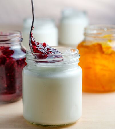Homemade yogurt with red berries marmalade spoon eating from a glass jar on a white wooden table. Healthy dairy probiotic breakfast.の写真素材