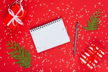 Holiday background , gift boxes with ribbon and bow and thuja twigs on a red background with glitter silver stars, open spiral notepad and pen, flat lay, top viewの写真素材