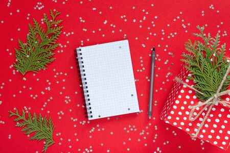 Holiday background , red gift box in polka dots with ribbon and bow and thuja twigs on a red background with glitter silver stars,open spiral notepad and pen, flat lay, top viewの写真素材
