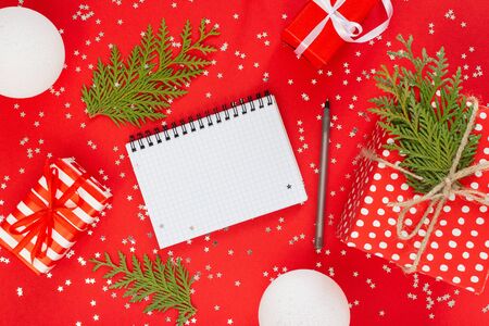 Holiday background , gift boxes with ribbon and bow and christmas balls and thuja twigs on a red background with glitter silver stars, open spiral notepad and pen, flat lay, top viewの写真素材
