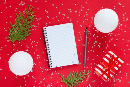 Holiday background , gift box with ribbon and bow and christmas balls and thuja twigs on a red background with glitter silver stars, open spiral notepad and pen, flat lay, top viewの写真素材
