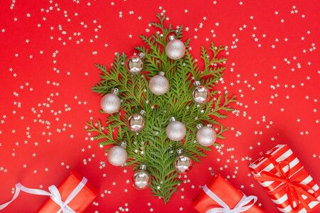 Decorative fir tree made of thuja twigs with Christmas balls, gift boxes on a red background with glitter silver stars, holiday backgroundの写真素材