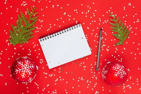 Holiday background , christmas balls with snowflakes and thuja twigs on a red background with glitter silver stars, open spiral notepad and pen, flat lay, top viewの写真素材