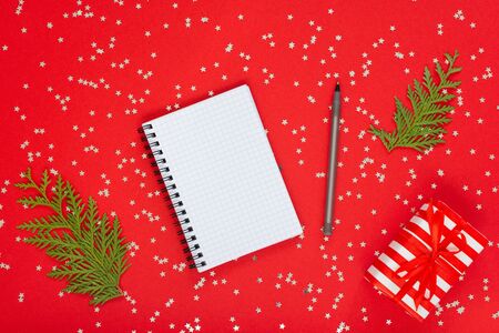 Holiday background , gift box with ribbon and bow on a red background with glitter silver stars, open spiral notepad and pen, flat lay, top viewの写真素材