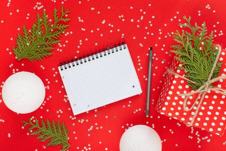 Holiday background , red gift box in polka dots with ribbon and bow and christmas balls and thuja twigs on a red background with glitter silver stars,open spiral notepad and pen, flat lay, top viewの写真素材