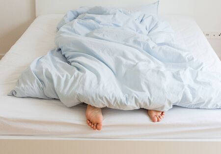 Part of the home or hotel interior, female legs peek out from under the covers, woman sleeping on a white bed with blue linens in the morningの写真素材
