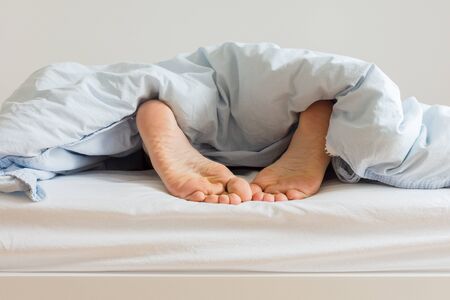 Part of the home or hotel interior, male legs peek out from under the covers, man sleeping on a white bed with blue linens in the morningの写真素材