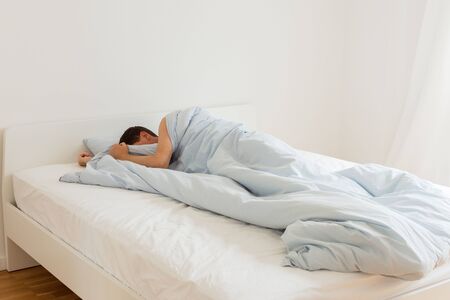 Part of the home or hotel interior, man sleeping on a white bed with blue linens in the morningの写真素材