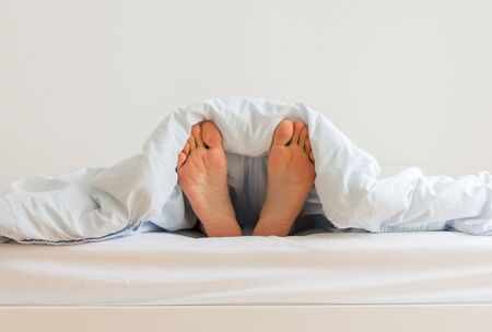 Part of the home or hotel interior, male legs peek out from under the covers, man sleeping on a white bed with blue linens in the morningの写真素材