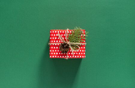 Holiday background , red gift box in polka dots and thuja twigs with christmas tree cone on green background, merry christmas and happy new year concept, flat lay, top viewの写真素材