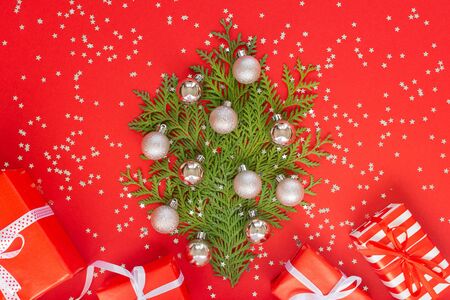 Decorative fir tree made of thuja twigs with Christmas balls, gift boxes on a red background with glitter silver stars, holiday backgroundの写真素材