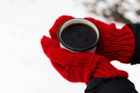 Female hands in red knitted mittens, in the hands of an iron mug with coffee or tea on a background of white snowの写真素材