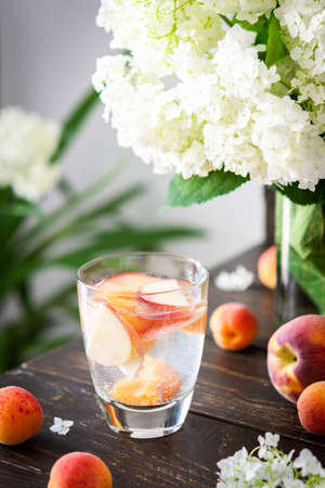 Refreshing drink with apricots and peaches, cold sparkling water with fruits, a vase of flowers viburnum opulus Roseum and a glass with a drinkの写真素材