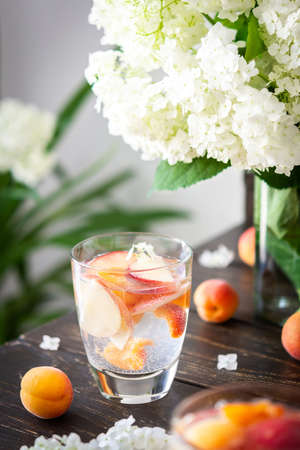 Refreshing drink with apricots and peaches, cold sparkling water with fruits, a vase of flowers viburnum opulus Roseum and a glass with a drinkの写真素材