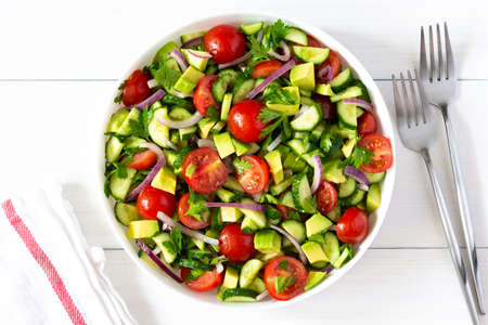Salad of fresh ripe tomato, onion, avocado, cucumber and parsley and olive oil, salad in a white plate with a fork and towel on a white wooden backgroundの写真素材