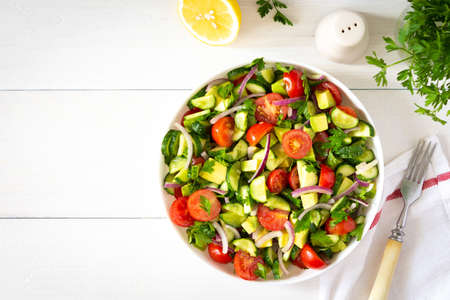 Salad of fresh ripe tomato, onion, avocado, cucumber and parsley and olive oil, salad in a white plate with a fork and towel on a white wooden backgroundの写真素材