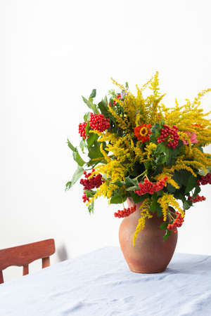 Beautiful bouquet of ragweeds, zinnia and branches of viburnum in a clay pot on a table against a white wallの写真素材