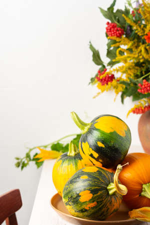 Beautiful bouquet of ragweeds, zinnia and branches of viburnum in a clay pot, fresh harvest of pumpkins on a table against a white wallの写真素材